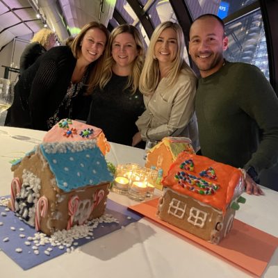 A group of four participants smiling and standing behind a table displaying three colorfully decorated gingerbread houses during a gingerbread house team building competition. The table is decorated with small lit candles, adding a cozy touch to the festive atmosphere. thumbnail