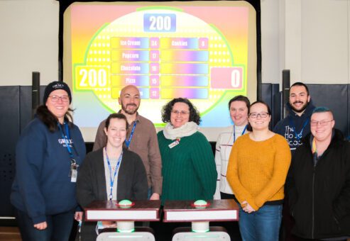 A group of eight participants smiling and standing together in front of a game show setup with a brightly colored game board in the background during the Corporate Feud game.