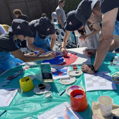 A group of participants in matching hats and aprons collaborate on painting a vibrant section of a mural during The Big Picture team building event. They work together, adding colors to the canvas under a sunny outdoor setting, with painting supplies and color wheels scattered on the table. thumbnail