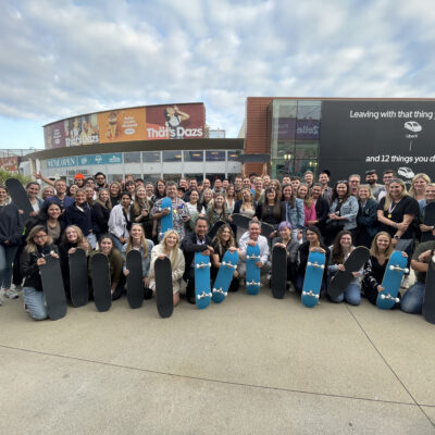 Large group of participants proudly display skateboards they built together during The Mystery Bus team building event, fostering teamwork and creativity. thumbnail