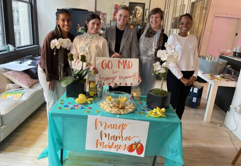 A team posing proudly behind their colorful 'Mango Mama Madness' station, showcasing their creativity in a fun and vibrant atmosphere during a team building event.