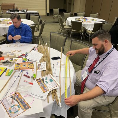 Two participants measure materials and work together to plan their bridge during the Bridging the Divide team building activity. thumbnail