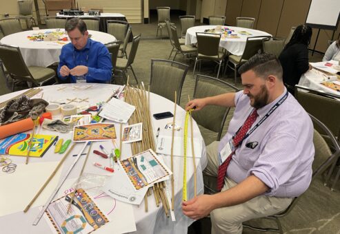 Two participants measure materials and work together to plan their bridge during the Bridging the Divide team building activity.
