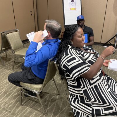 Two participants engage in a collaborative communication exercise, sitting back-to-back as part of the Bridging the Divide team building activity. thumbnail