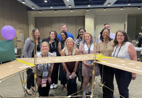 A team poses proudly behind their completed bridge structure during the Bridging the Divide team building activity.