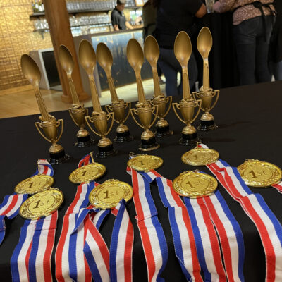 A table displays several gold trophies in the shape of large spoons and medals with '1st' on them. The medals are attached to red, white, and blue ribbons. The trophies and medals are for the culinary team building winners. thumbnail