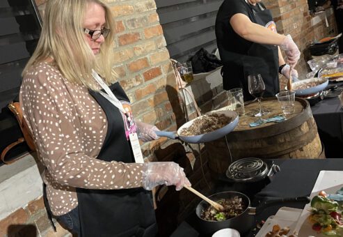 Two women participating in a team building chili cook-off. Both are focused on preparing food at their respective stations. The woman on the left is stirring ingredients in a bowl, wearing a brown polka-dotted cardigan over her apron. The woman on the right is cooking something on a skillet while wearing a black shirt and apron.