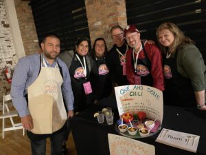 A group of six participants posing at a team building chili cook-off event behind their table. The table displays a sign reading 'One and Done Chili' with a description and a tray of chili servings garnished with toppings. The participants are smiling and wearing 'Chili Cook-Off' aprons.