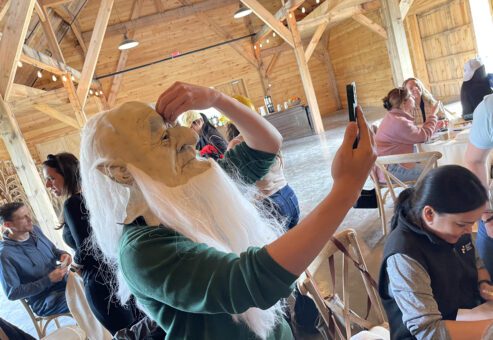 Person wearing a long white-bearded mask taking a selfie with a smartphone, preparing for the Corporate Film Festival challenge.