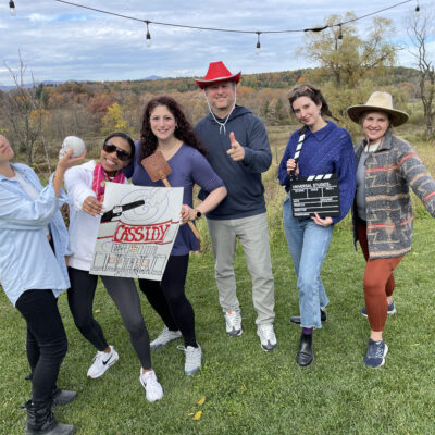 A group of six people pose outdoors, holding various props like a film clapperboard, a poster, and a red cowboy hat, participating in a creative team building activity themed around film production and storytelling. thumbnail