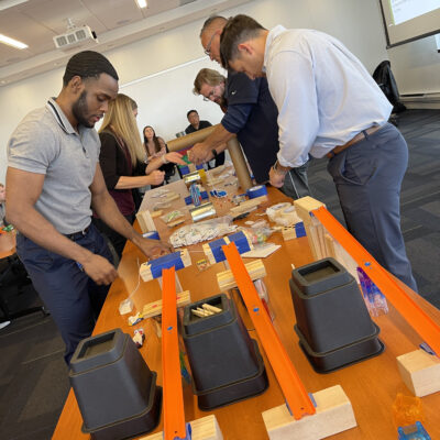 Team members collaborating during the Domino Effect team building event, working with various materials and components on a table to create complex contraptions. thumbnail