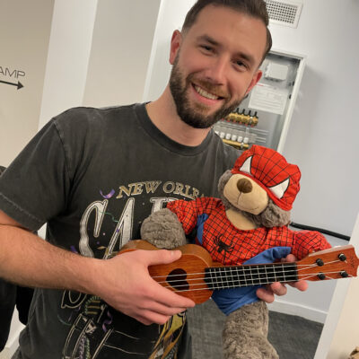 A smiling man holds a teddy bear dressed in a Spider-Man costume while also holding a small ukulele. The man is participating in a Toys for Tykes team building event, proudly showcasing the decorated toy ready to be donated to a child. thumbnail