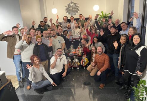 A large group of people smiling and waving for a group photo. They are gathered around a collection of decorated toys and gifts, assembled during a CSR team building program.