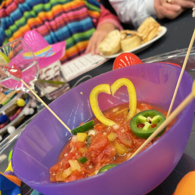 A colorful bowl of salsa garnished with heart-shaped yellow peppers and jalapeños at a Salsa Showdown team building event, with participants wearing vibrant striped outfits. thumbnail