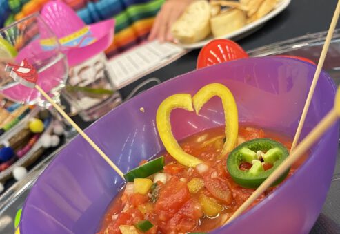 A colorful bowl of salsa garnished with heart-shaped yellow peppers and jalapeños at a Salsa Showdown team building event, with participants wearing vibrant striped outfits.