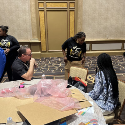 A group of people seated around a table in a large conference room working on a team building project. The table is covered with various crafting supplies, including bubble wrap, cardboard, markers, and a sketchpad. One man is seated, appearing to work or think while another person, seated next to him, is writing on paper. A woman in a black t-shirt is standing near the table, holding a piece of material or cardboard involved in the activity. The participants are engaging in a creative task, designing a garment together. thumbnail