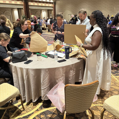 Participants at a team building event are gathered around a round table, actively engaging in a design project. They are cutting and folding pieces of cardboard and other materials as part of a creative challenge. A woman in a white dress stands next to the table, holding a piece of cardboard, while others around her are focused on their tasks. The room is filled with other groups working on similar projects. thumbnail