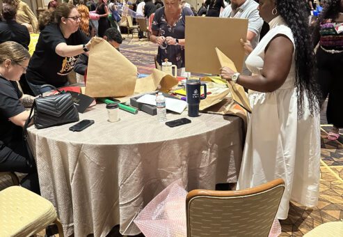Participants at a team building event are gathered around a round table, actively engaging in a design project. They are cutting and folding pieces of cardboard and other materials as part of a creative challenge. A woman in a white dress stands next to the table, holding a piece of cardboard, while others around her are focused on their tasks. The room is filled with other groups working on similar projects.
