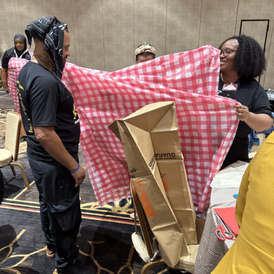 Two participants at a team building event stand, with one holding a large red and white checkered fabric, while the other appears to be modeling it or assisting in its setup. Cardboard and other recyclable materials are visible, being used to create an outfit. thumbnail