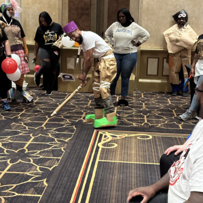 A group of people participate in a fun team building event inspired by a fashion runway show. The focus is on one individual wearing a creative outfit made from paper and other recyclable materials, including a purple hat, brown pants, and neon green shoes. The individual is in an action pose, holding a stick as if preparing for a playful activity. Several other participants, also dressed in quirky, recyclable material outfits, stand around watching. thumbnail