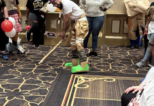 A group of people participate in a fun team building event inspired by a fashion runway show. The focus is on one individual wearing a creative outfit made from paper and other recyclable materials, including a purple hat, brown pants, and neon green shoes. The individual is in an action pose, holding a stick as if preparing for a playful activity. Several other participants, also dressed in quirky, recyclable material outfits, stand around watching.