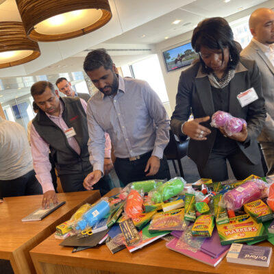 Group of professionals sorting through school supplies like markers, crayons, and notebooks on a table, preparing backpacks for donation at a Tools for Schools CSR team building event. thumbnail