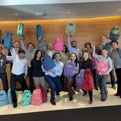 A group of smiling participants proudly holding up backpacks filled with school supplies during a Tools for Schools CSR team building event. thumbnail
