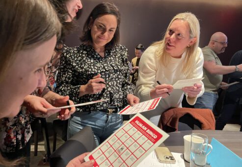 Group of colleagues engaging in a fun 'Human Bingo' team building activity, marking their bingo cards and interacting with one another.