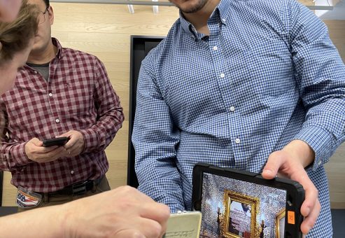 Team member holding a tablet showing a virtual crime scene as others examine a small evidence item during the Corporate CSI team building event.