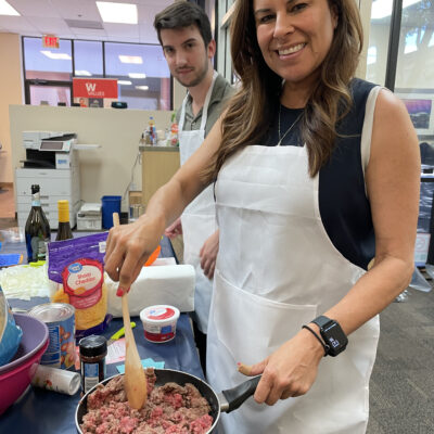 A woman and a man participate in a culinary team building event. The woman is smiling as she stirs a pan filled with ingredients while wearing a white apron. Various food items like cheese and sour cream are visible on the table in front of them. The man behind her is also wearing an apron, standing by the food preparation area. thumbnail
