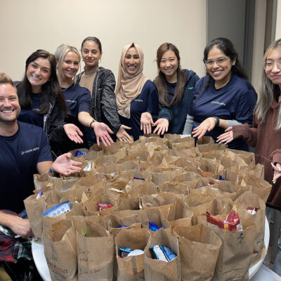 A group of eight smiling individuals stand around a table filled with brown paper bags, likely packed with food or care items. They are proudly showing off their work, extending their hands toward the bags. Everyone in the group appears happy and engaged in this Do Good Bus team building event. thumbnail
