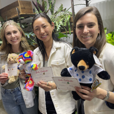 Three women smiling and holding colorful stuffed animals and birth certificates they created during a corporate charitable team building event called The Donation Station. thumbnail