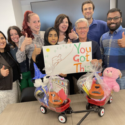 A group of eight people stand together smiling and giving thumbs up around two small Radio Flyer wagons filled with teddy bears and toys. One person is holding a handmade sign that says 'You Got This!' with a colorful drawing. The team appears happy and proud, showcasing their contributions during a team building activity focused on charity and giving back. thumbnail