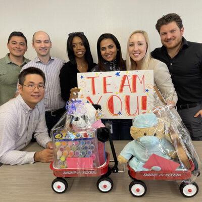 A team of six people proudly showcases their completed toy wagons filled with stuffed animals, arts and crafts supplies, and a sign that reads 'TEAM YOU!' as part of a Toys for Tykes team building event. thumbnail