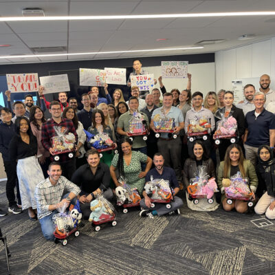 A large group of people smiling and posing together in a modern office space. They are holding and sitting with small red Radio Flyer wagons filled with various toys, teddy bears, and musical instruments, all wrapped in gift wrap. Behind them, some participants are holding up hand-drawn signs with motivational messages like 'You Got This!' This photo captures the conclusion of the Toys for Tykes ‘Arts & Music’ CSR team building event. thumbnail