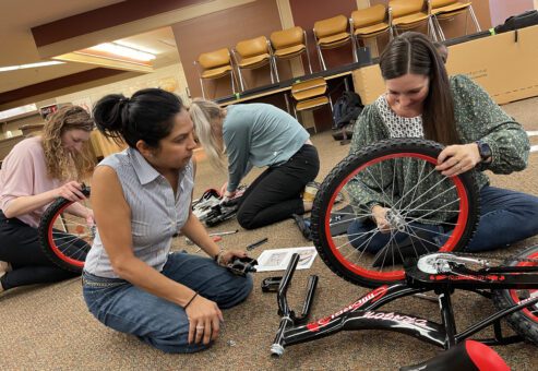 Four team members work together on the floor to assemble bikes during the Charity Bike Build event. They are focused on attaching wheels and following instructions, demonstrating collaboration and problem-solving as part of this hands-on, team building activity.