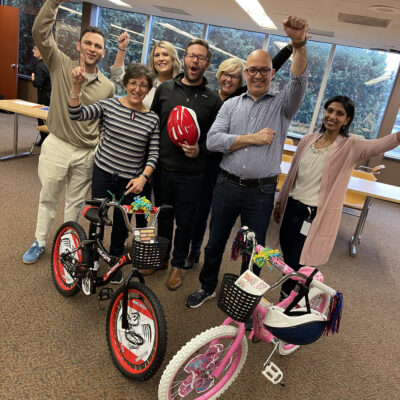 A group of seven participants cheer enthusiastically while standing behind two completed bikes during a Charity Bike Build event. thumbnail
