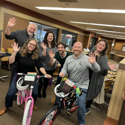 A group of six people smile and wave after successfully assembling two bicycles—one pink and one black—during a Charity Bike Build event. The team members stand proudly next to the bikes they built, showcasing their collaboration and effort. thumbnail