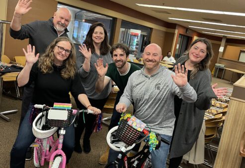 A group of six people smile and wave after successfully assembling two bicycles—one pink and one black—during a Charity Bike Build event. The team members stand proudly next to the bikes they built, showcasing their collaboration and effort.