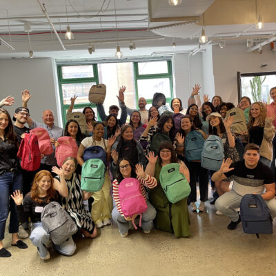 Group of enthusiastic participants holding colorful backpacks filled with school supplies, celebrating the successful completion of the Tools for Schools charity event in a bright and modern space. thumbnail