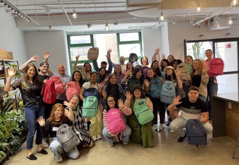 Group of enthusiastic participants holding colorful backpacks filled with school supplies, celebrating the successful completion of the Tools for Schools charity event in a bright and modern space.