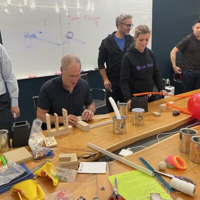 Team members collaborating and building creative contraptions during the Domino Effect team building event, surrounded by various building materials and tools. thumbnail