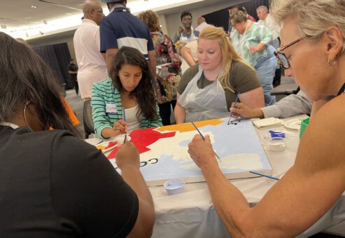 A group of participants sits around a table during The Big Picture team building event, painting a canvas section with various colors. They focus on filling in their assigned part of the mural, collaborating to complete the larger artwork.