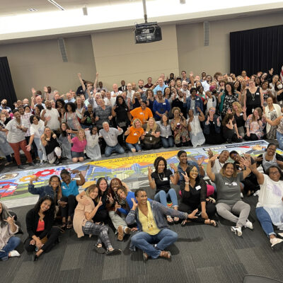 A large group of participants celebrates completing a collaborative mural during The Big Picture team building event, showcasing teamwork and creative achievement. thumbnail