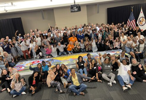 A large group of participants celebrates completing a collaborative mural during The Big Picture team building event, showcasing teamwork and creative achievement.