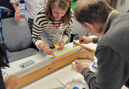 Participants collaborate to paint and decorate a wooden Little Free Library mounting post during a team building event. Each team contributes their creativity to create a cohesive design, fostering teamwork and connection while working toward the shared goal of building a community library.