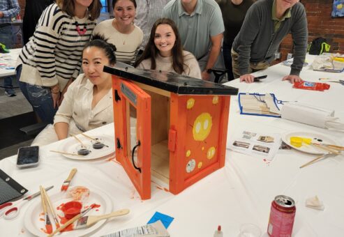 Participants gather around a table to mix and paint with vibrant colors during a creative team building activity. The hands-on session encourages collaboration and artistic expression as teams work together to design elements for a Little Free Library, promoting teamwork and community engagement.