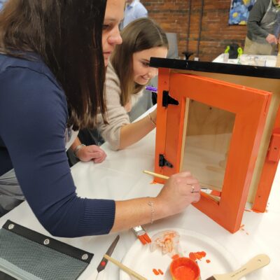 Participants focus on painting a Little Free Library door with bright orange during a collaborative team building event. The hands-on activity fosters creativity and teamwork as teams work together to build and decorate the library for community use. thumbnail