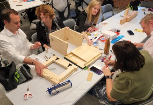 Teams collaborate to assemble a Little Free Library during a hands-on team building event. Participants work together to construct the wooden structure, following instructions and dividing tasks to create a functional and beautifully decorated library for the community.