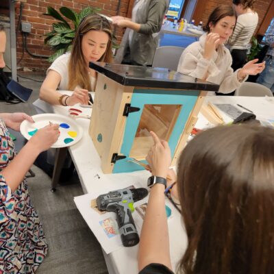 Participants focus on painting a Little Free Library door with bright orange during a collaborative team building event. The hands-on activity fosters creativity and teamwork as teams work together to build and decorate the library for community use. thumbnail